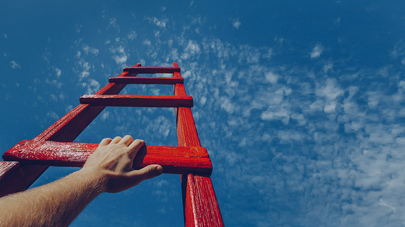 A male hand holds onto the crossbar of a red wooden staircase leading to the blue sky
